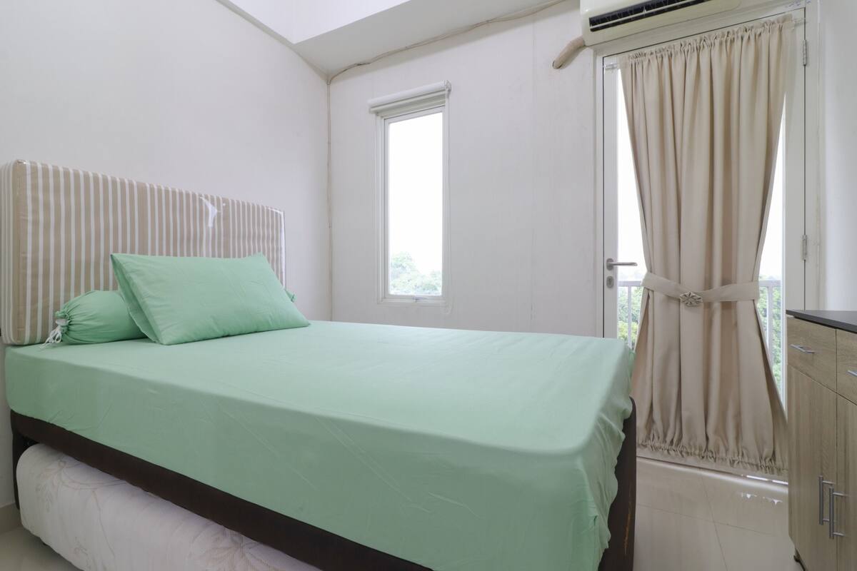 A single bed with a green bedspread and two pillows, a window with a view, and a door with curtains.