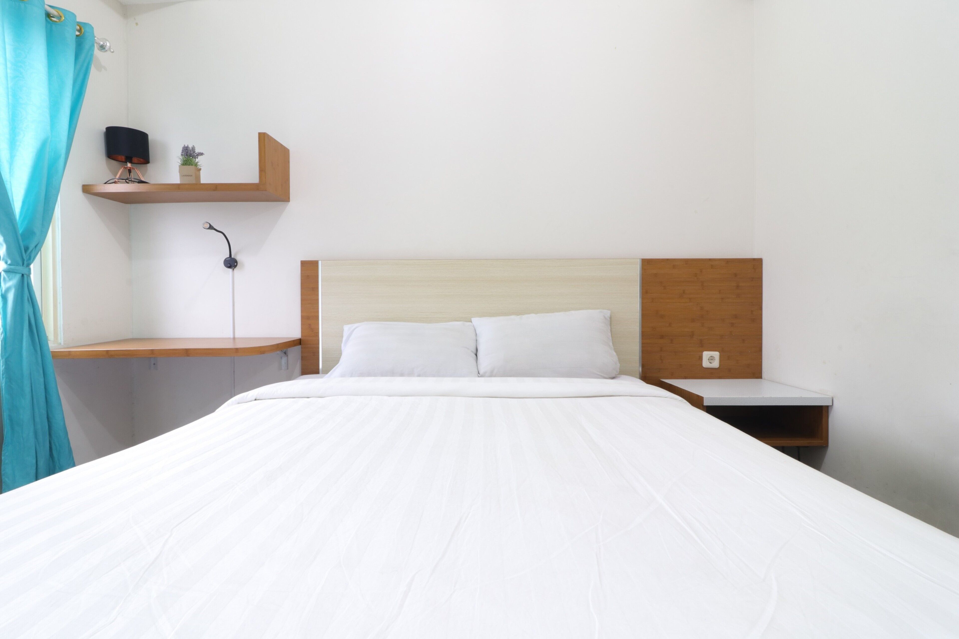 A neatly made bed with white linens, a wooden headboard, and a small shelf with a lamp and a plant.