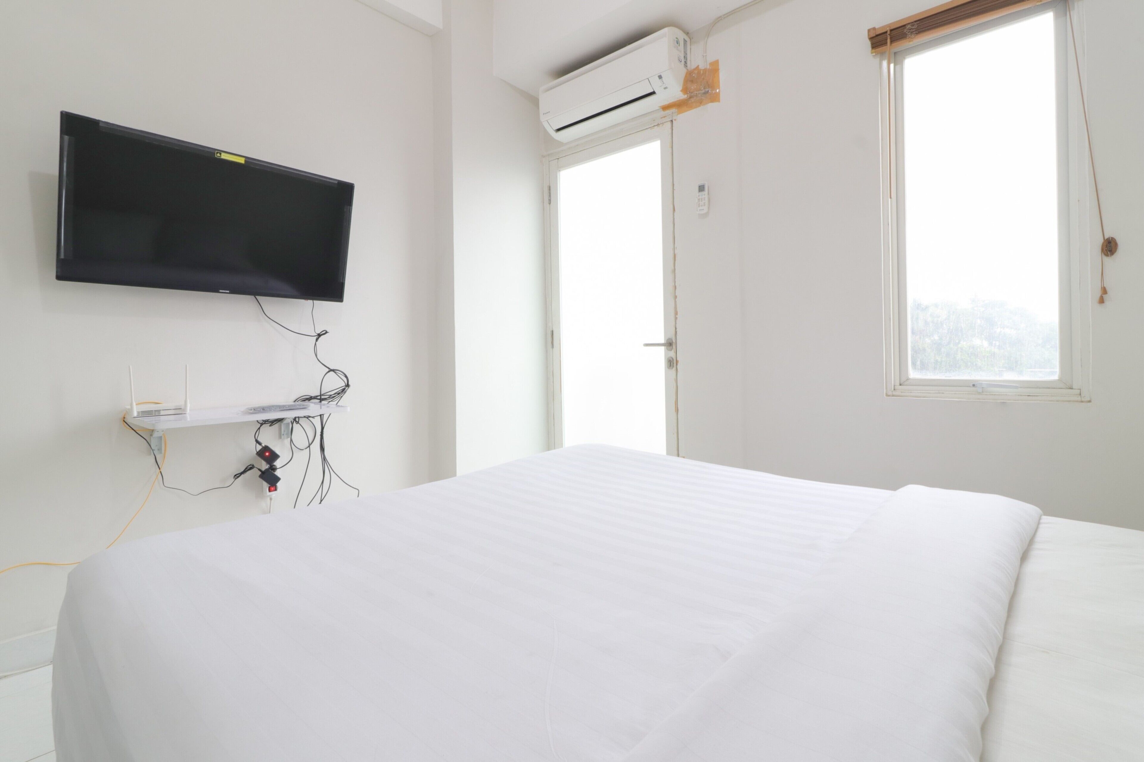 A bedroom with a bed, a mounted TV, an air conditioning unit, and a window with blinds.