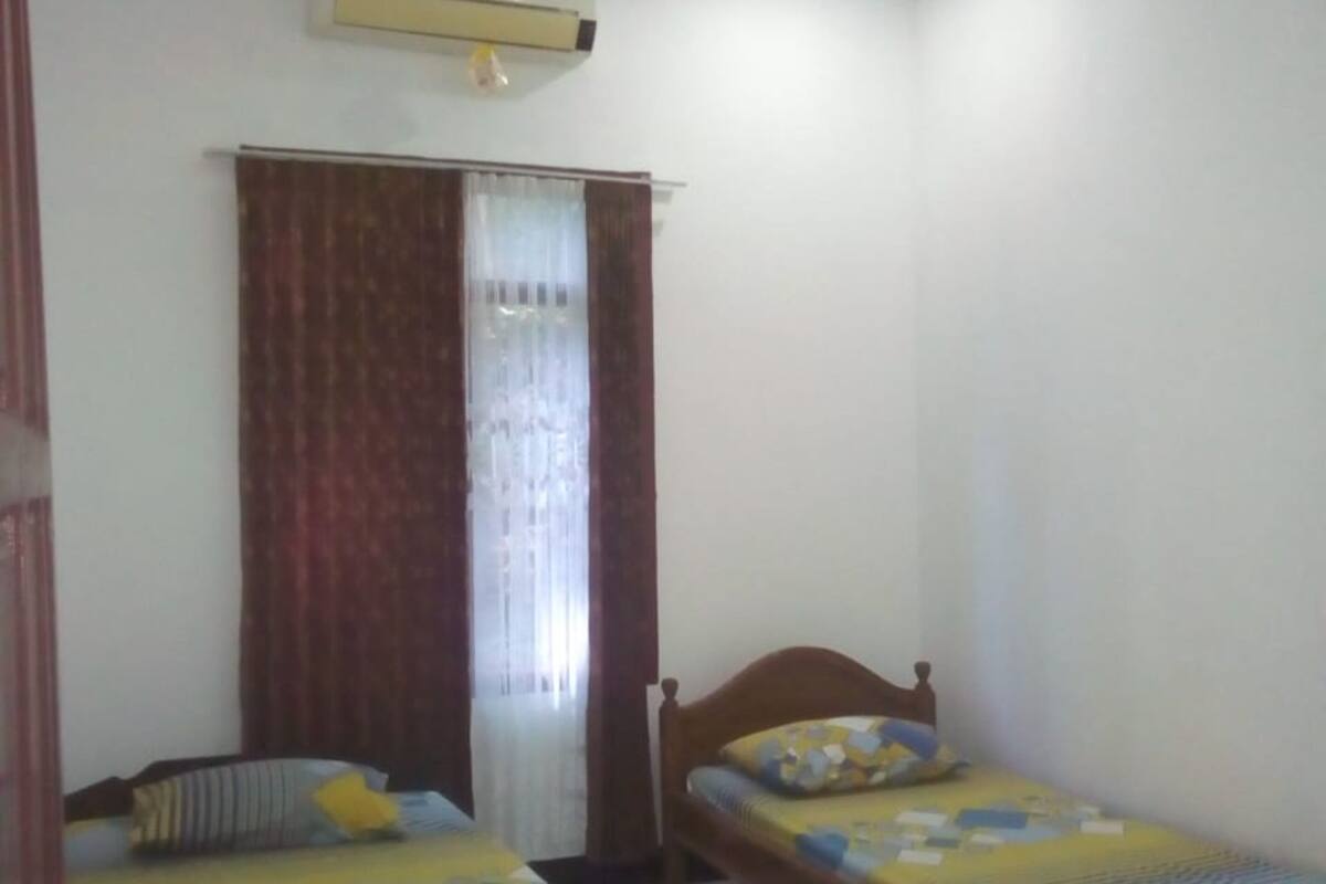 Ein Zimmer mit zwei Einzelbetten, einem fensterbelaubten Fenster, einer Klimaanlage und einem Fliesenboden.