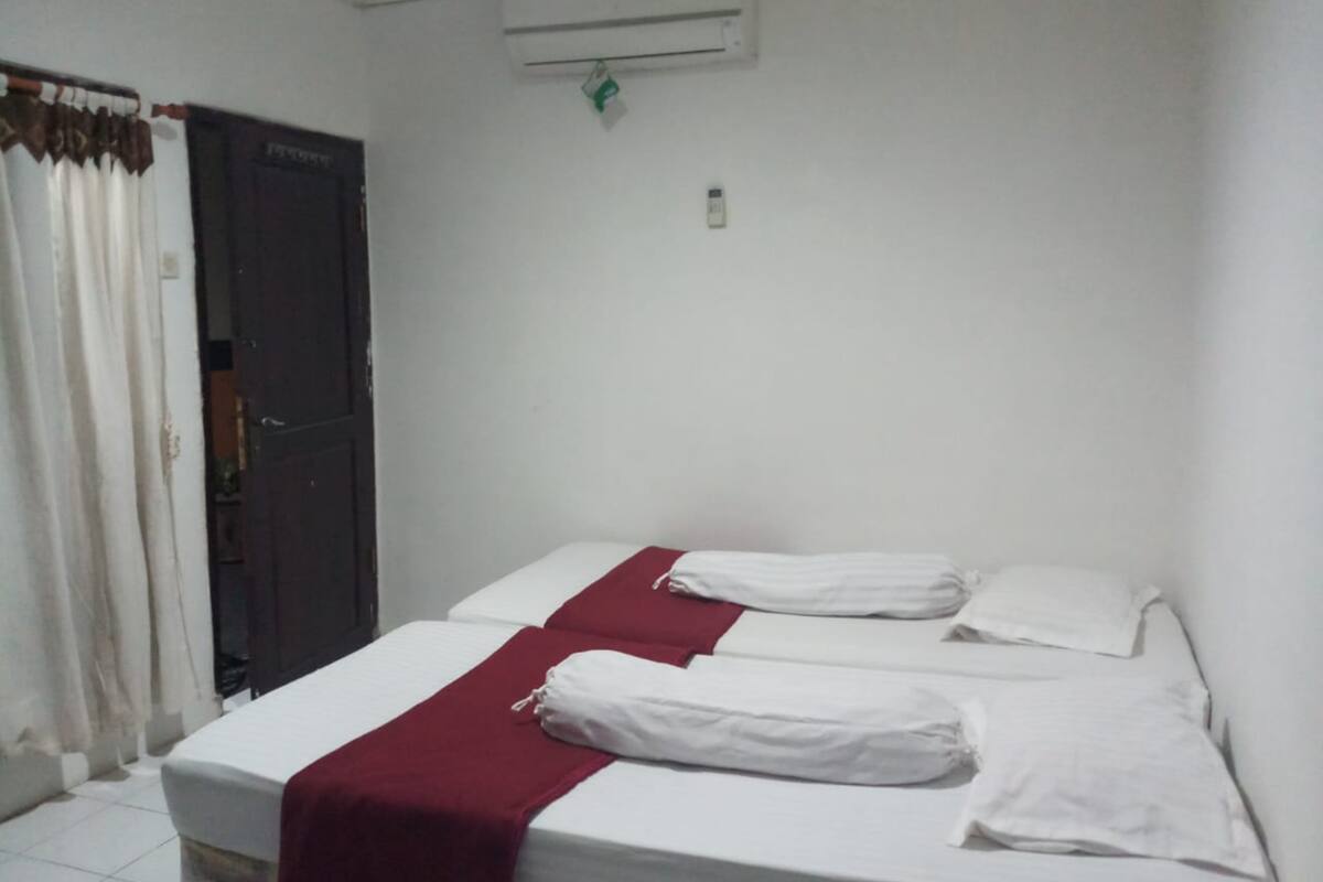 A single bed with white bedding and a red blanket, a closed dark-colored door, and an air conditioning unit on the wall.