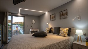 A bedroom with a large bed, bedside table, and a view of a balcony with plants. - Mini Bella Gonia (Rethymno)