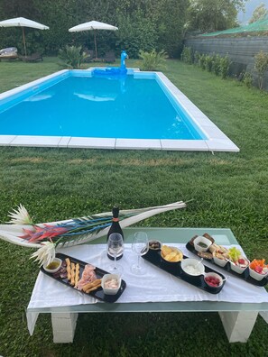 Zona de piscina amb taula plena d'aperitius i begudes, envoltada d'un verdós prat i una piscina blava.