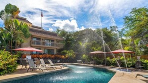 Ein Schwimmbecken mit Wasserfall, umgeben von Liegestühlen und Sonnenschirmen, mit einem Gebäude im Hintergrund.