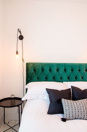 A bed with a green tufted headboard, white bedding, and multiple pillows. There is a small round table with a glass on it, and a wall-mounted light fixture. - Charming Boutique House – Stylish Stay Just Minutes from Margate Beach (Kent)