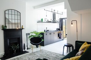 Ein modernes Wohnzimmer mit Kamin, einem schwarzen Sessel, einer grünen Couch und einer Küche im Hintergrund.