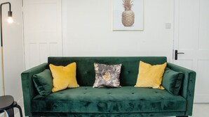 Un sofá de terciopelo verde con tres cojines, una obra de arte con forma de piña enmarcada en la pared y una lámpara de pie.
