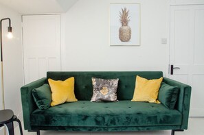 A green velvet sofa with three cushions, a framed pineapple artwork on the wall, and a floor lamp. - Charming Boutique House – Stylish Stay Just Minutes from Margate Beach (Kent)