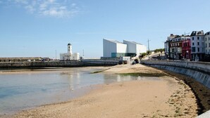 Zona costera con playa arenosa, un edificio moderno y un faro.