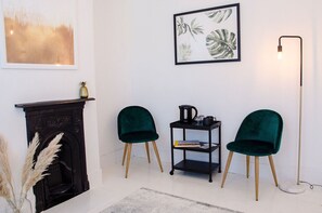 A room with a fireplace, two green chairs, a black side table with a kettle, a framed plant, and a floor lamp. - Charming Boutique House – Stylish Stay Just Minutes from Margate Beach (Kent)