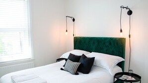 Una cama bien tendida con sábanas blancas y varias almohadas, un cabecero tapizado en verde y dos lámparas de noche con bombillas expuestas.