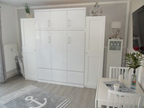 Ein Zimmer mit einem weißen Kleiderschrank, einem Esstisch mit einer Vase und einer Flasche sowie einem Heizkörper.