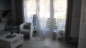 A living room with a white sofa, a white armchair, a small table with a plant, and a view of a garden through sheer curtains with anchor designs.