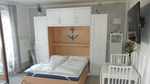 A bedroom with a bed, a desk, and a chair. There is a window with curtains, a radiator, and a plant on the wall.