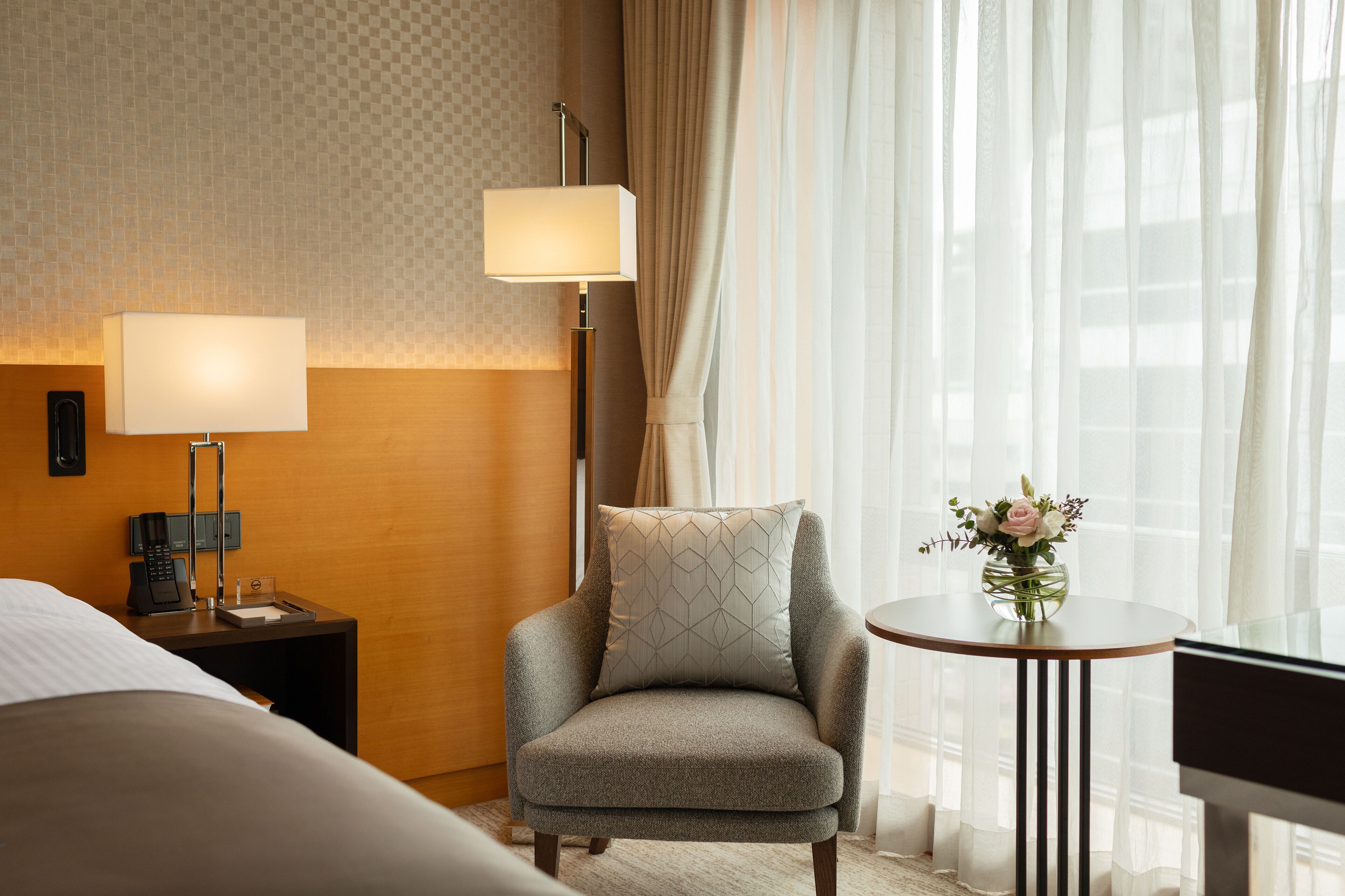 A hotel room with a bed, a chair with a patterned pillow, a bedside table with a lamp, a small table with a vase of flowers, and a window with sheer curtains.