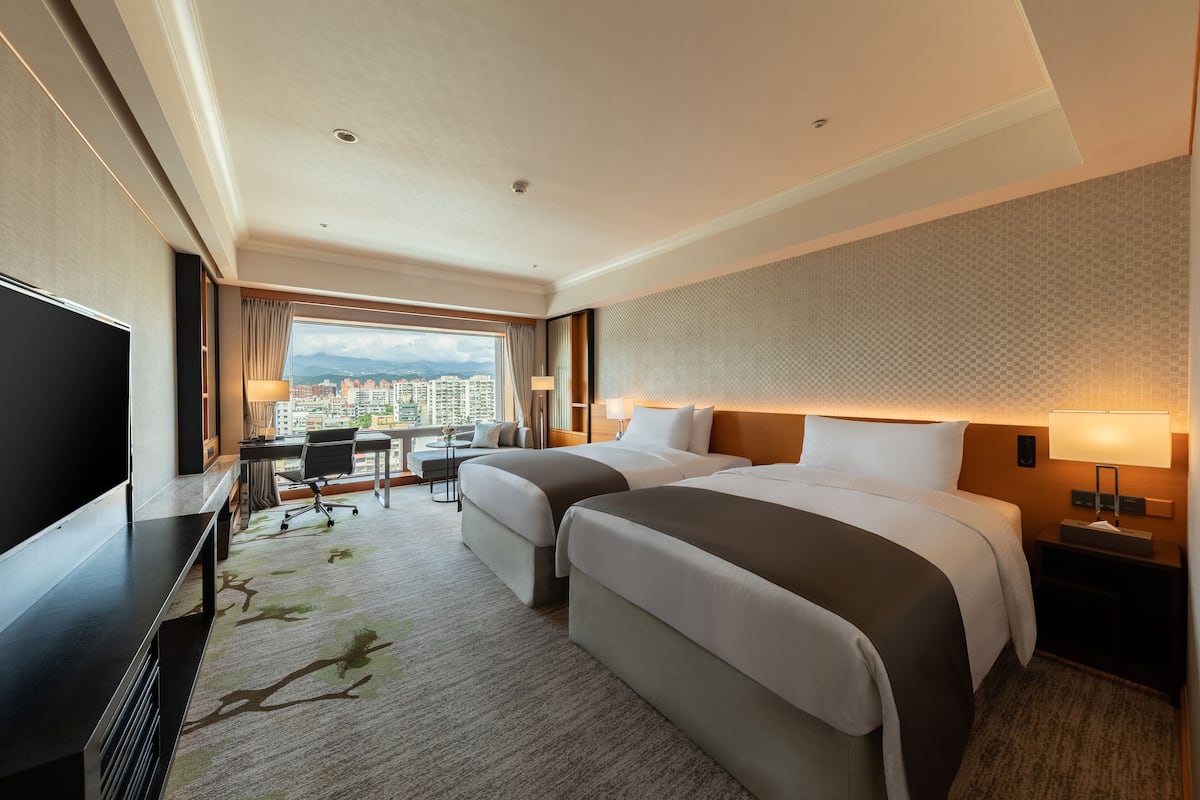 A hotel room with two beds, a desk, a TV, and a view of the city.