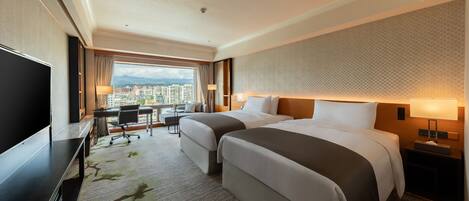 A hotel room with two beds, a desk, a TV, and a view of the city.