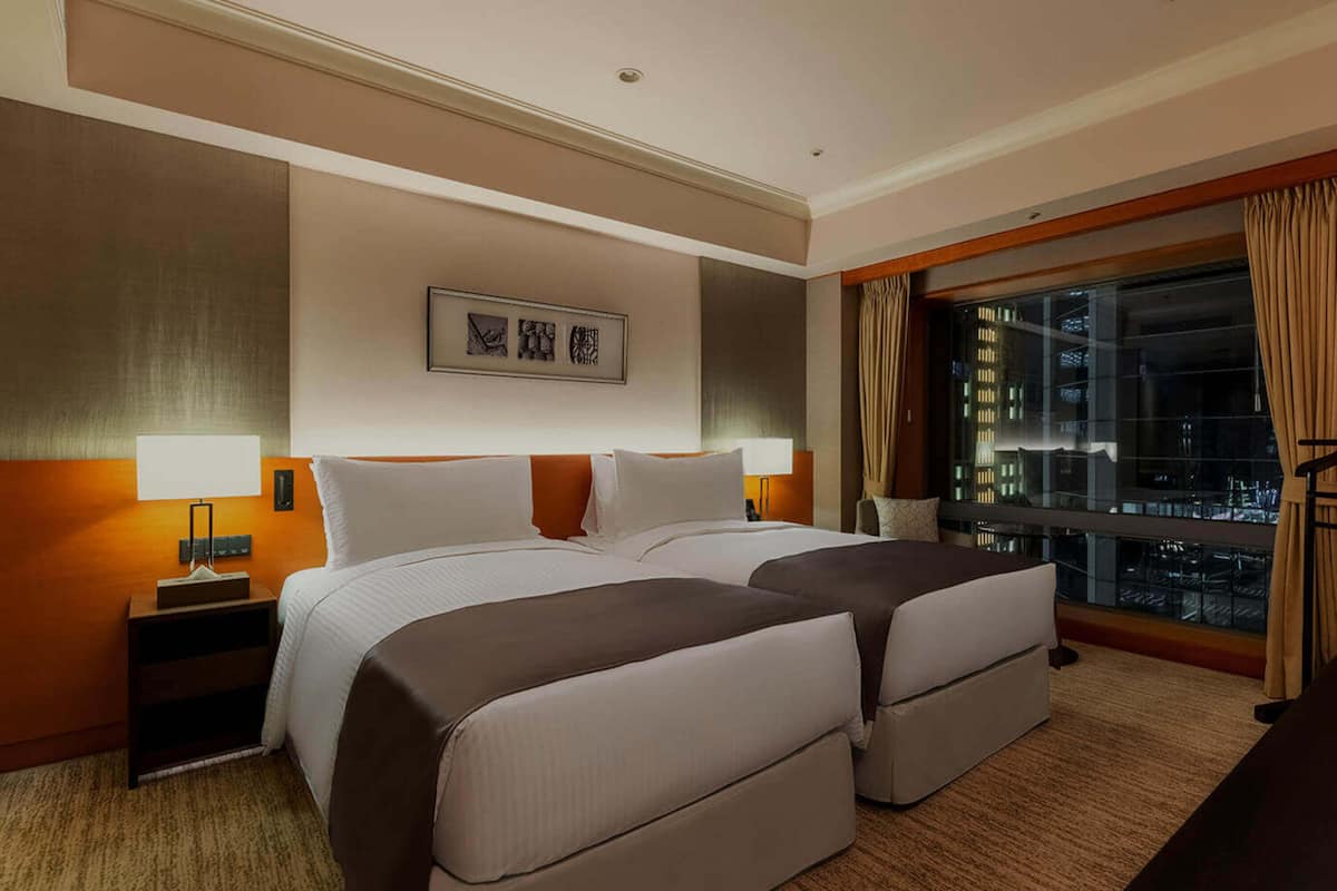 A hotel room with two beds, a nightstand, a lamp, a window with curtains, and a city view.