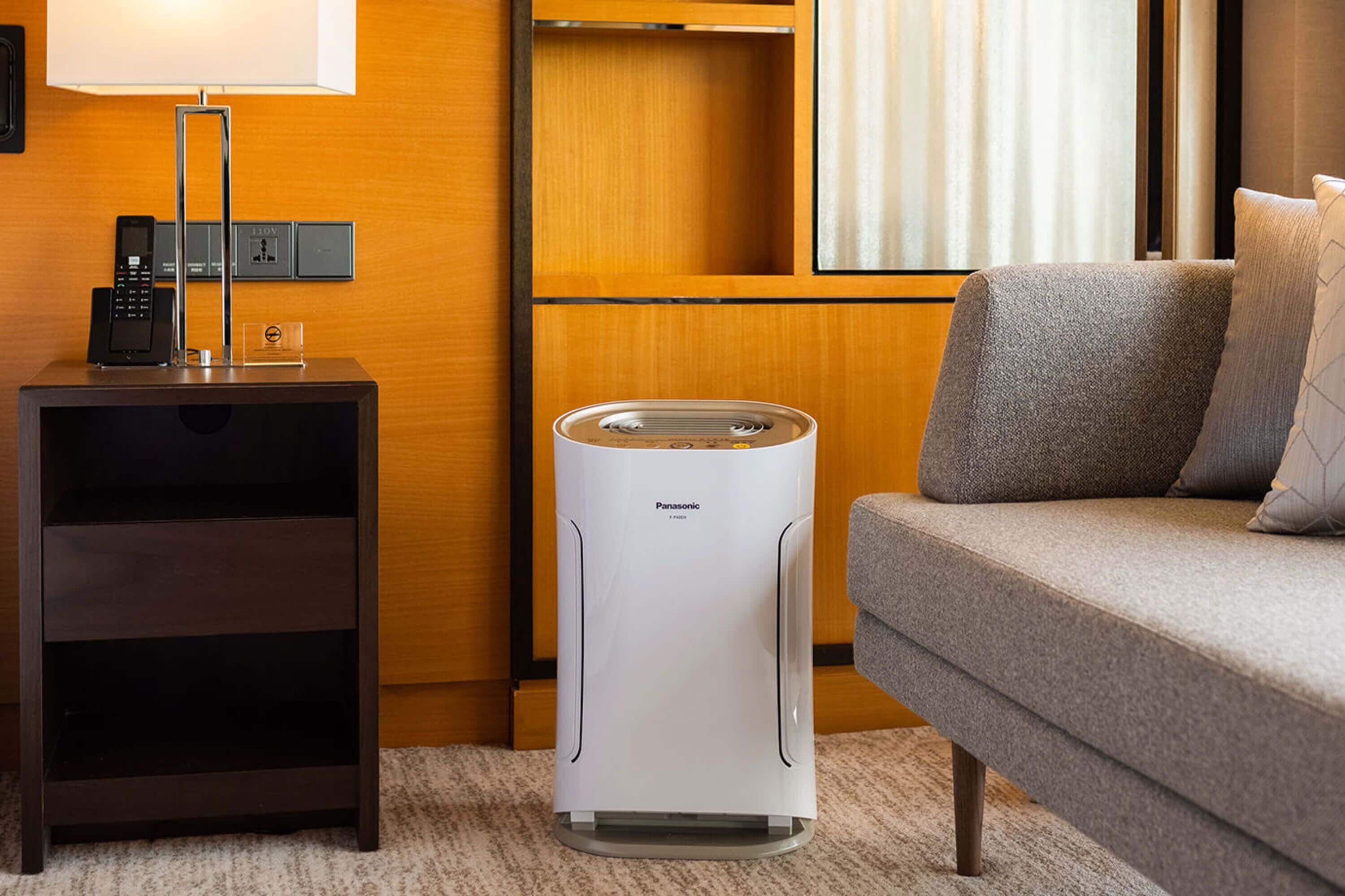 A hotel room with a bed, a bedside table with a lamp, a control panel, and a white air purifier.