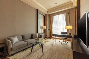 Een hotelkamer met een bank, een glazen salontafel, een bureau met stoel, een televisie en een raam met gordijnen.