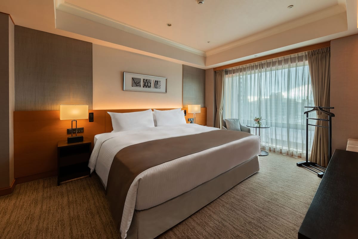 A hotel room with a large bed, bedside lamps, a small table with a vase, a chair, and a view of the city through sheer curtains.