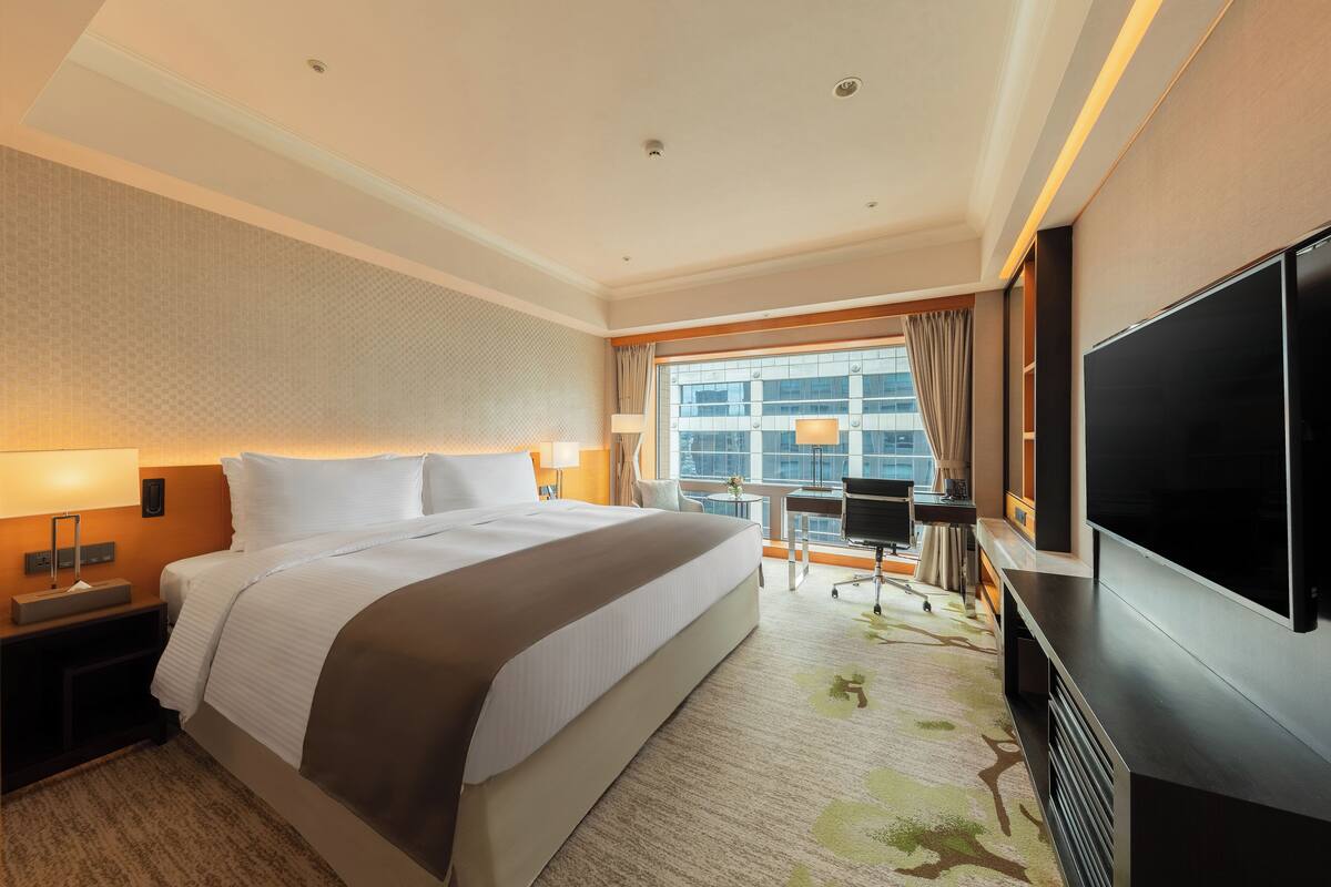 A hotel room with a large bed, a flat-screen TV, a desk with a chair, and a view of the city through the window.