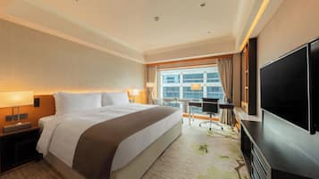 A hotel room with a large bed, a flat-screen TV, a desk with a chair, and a view of the city through the window.