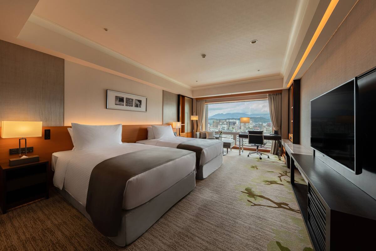 A hotel room with two beds, a desk, a TV, and a city view.