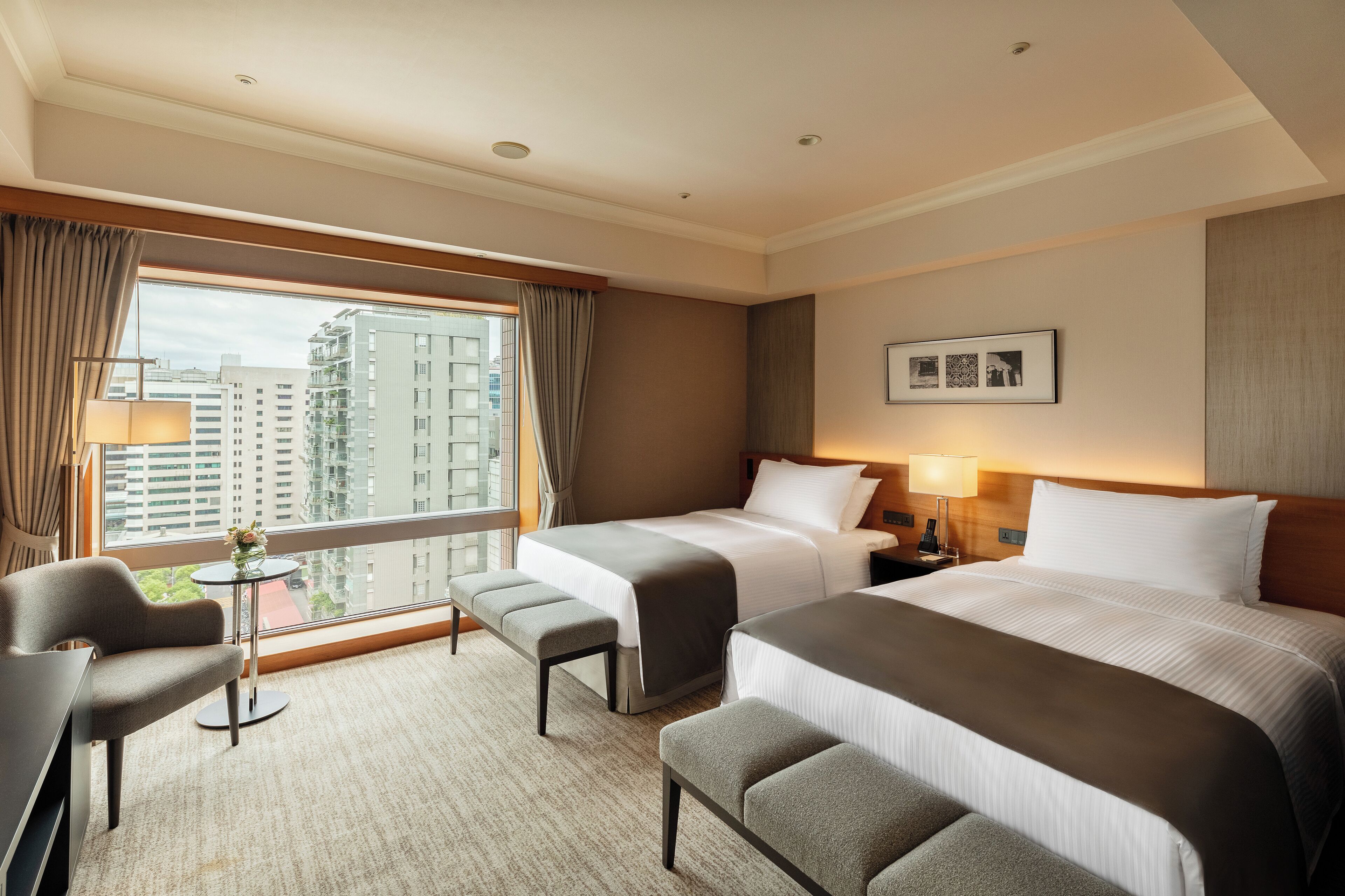 A hotel room with two beds, a desk, a chair, a small table, and a window with a view of buildings.