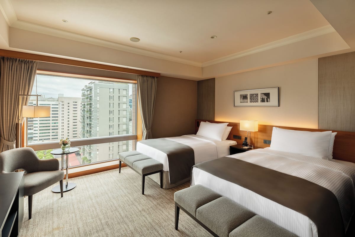 A hotel room with two beds, a desk, a chair, a small table, and a window with a view of buildings.