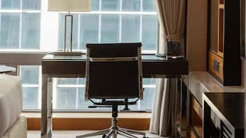A hotel room with a desk, chair, lamp, and a large window with curtains.