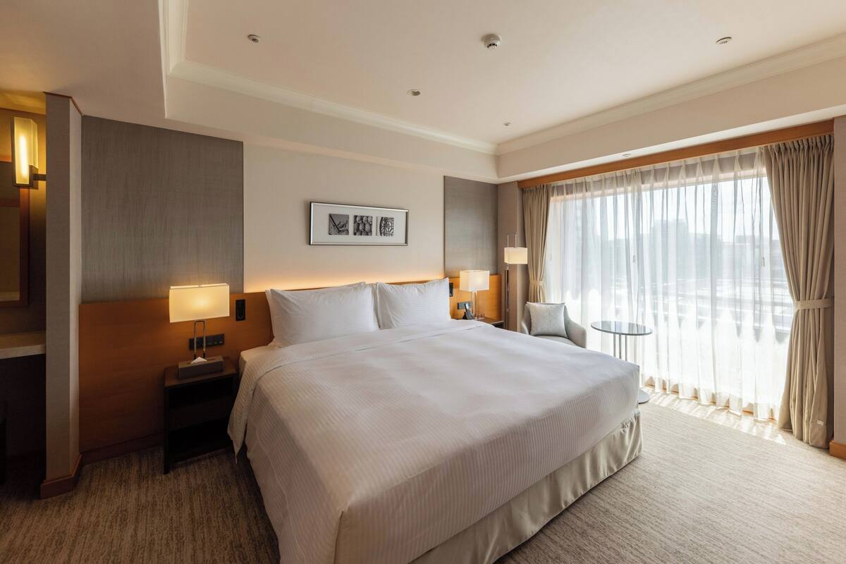 A hotel room with a large bed, two bedside tables with lamps, a small round table, a chair, and a large window with curtains.