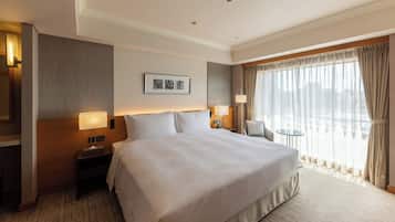 A hotel room with a large bed, two bedside tables with lamps, a small round table, a chair, and a large window with curtains.