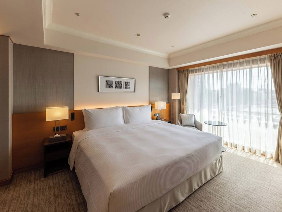 A hotel room with a large bed, two bedside tables with lamps, a small round table, a chair, and a large window with curtains.