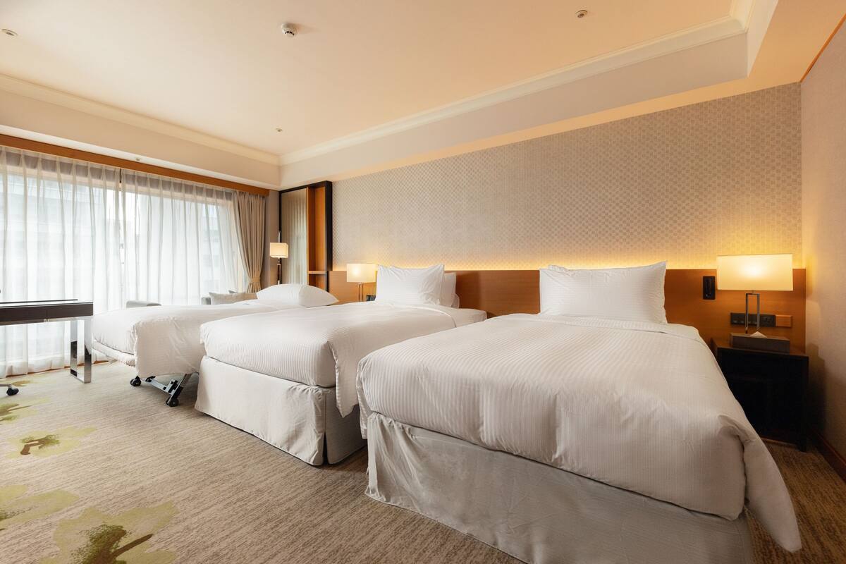 A hotel room with two beds, a desk, and a large window with curtains.