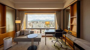 A hotel room with a sofa, a small round table, a desk, and a chair. There is a large window with a city view and mountains in the distance. - Hotel Metropolitan Premier Taipei (Taipei)