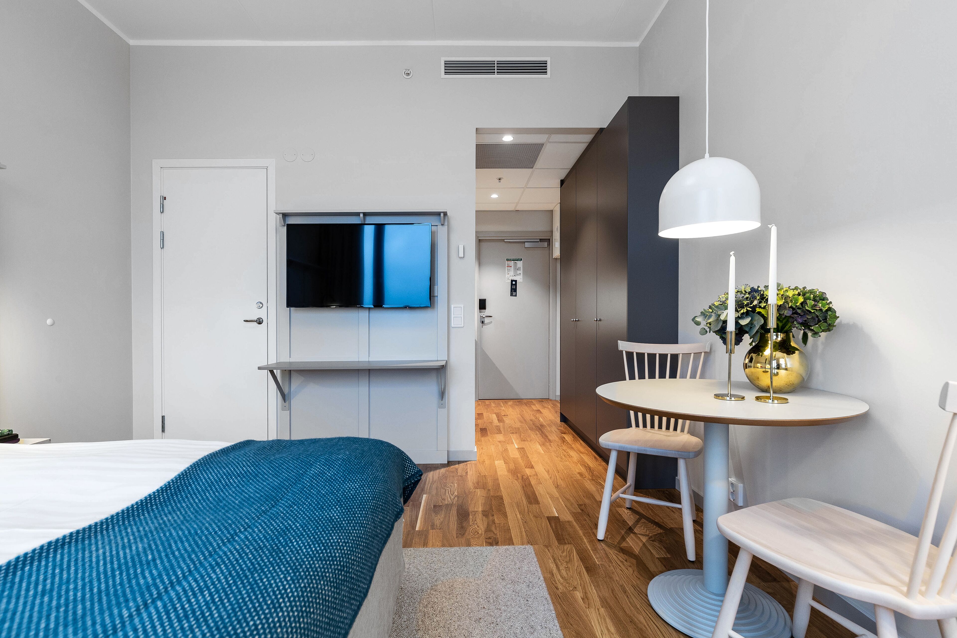 Una habitación de hotel moderna con una cama, un televisor fijado a la pared, una pequeña mesa de comedor con sillas y una planta en maceta.