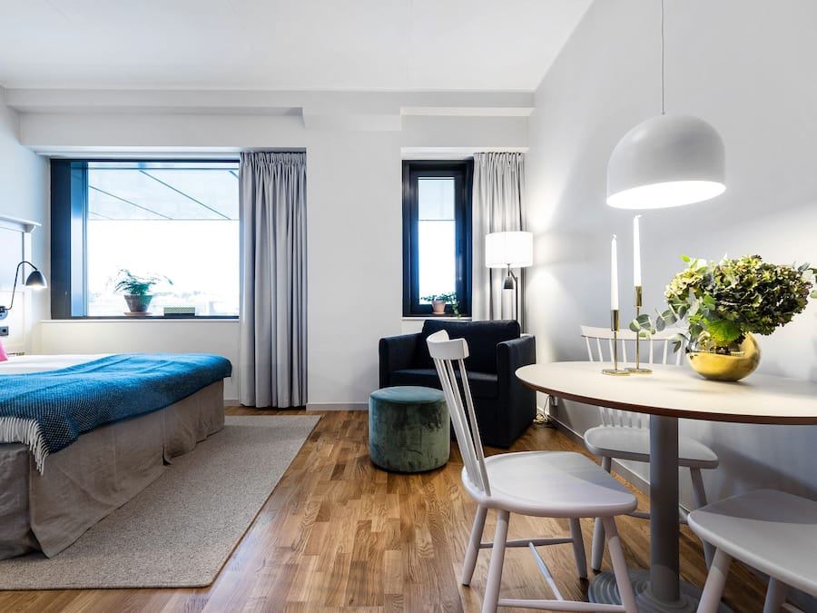 Moderni hotellihuone, jossa on sänky, pyöreä ruokapöytä, pieni pyöreä sivupöytä, tuoli, ruukkukasvi ja ikkuna, jossa on verhot.