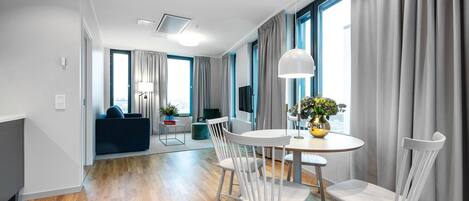 A modern hotel room with a dining area, wooden flooring, and large windows with curtains.