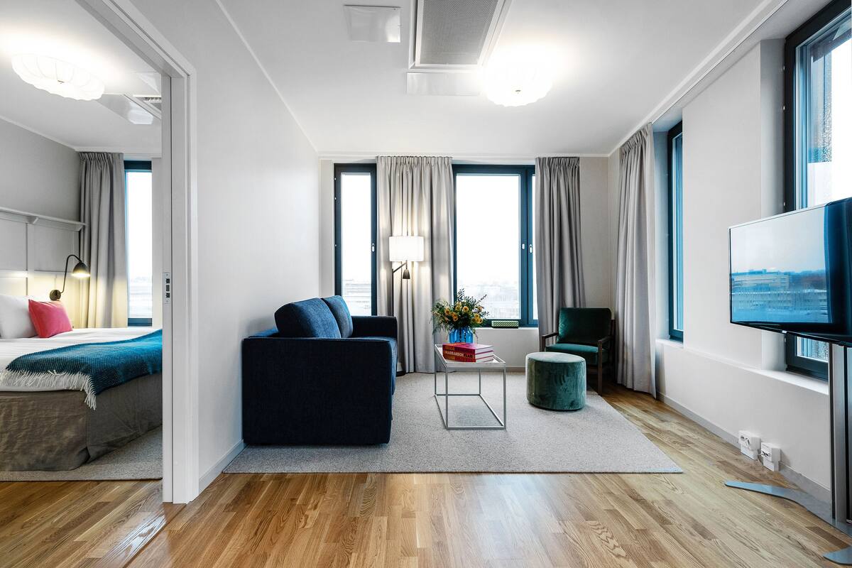 Una habitación de hotel moderna con una cama grande, un sofá oscuro, una mesita de noche con l ámpara, un televisor y vistas a la ciudad a través de amplios ventanales.