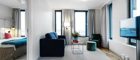 A modern hotel room with a large bed, a dark sofa, a small table with a lamp, a TV, and a view of the city through large windows.