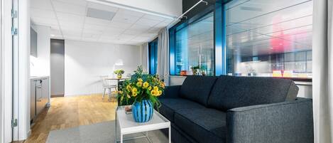 A modern office space with a grey sofa, a white table with a vase of flowers, and a large window with blue curtains.
