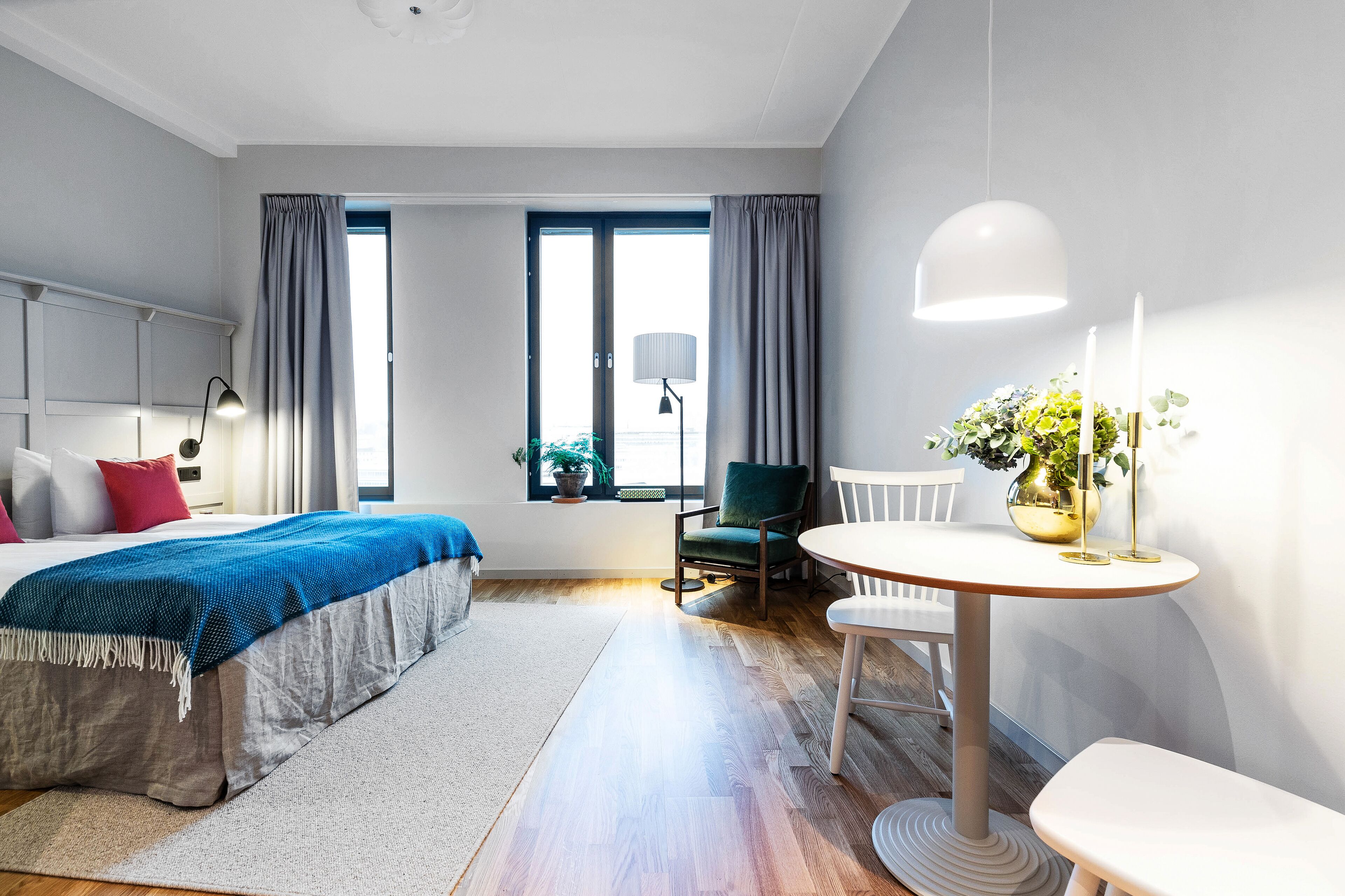 A modern hotel room with a large bed, a round dining table, a chair, a small table with candles, and a window with curtains.