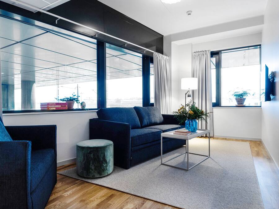A modern living room with a dark blue sofa, a matching armchair, a small round ottoman, a coffee table, and a floor lamp. Large windows with curtains provide natural light and views.