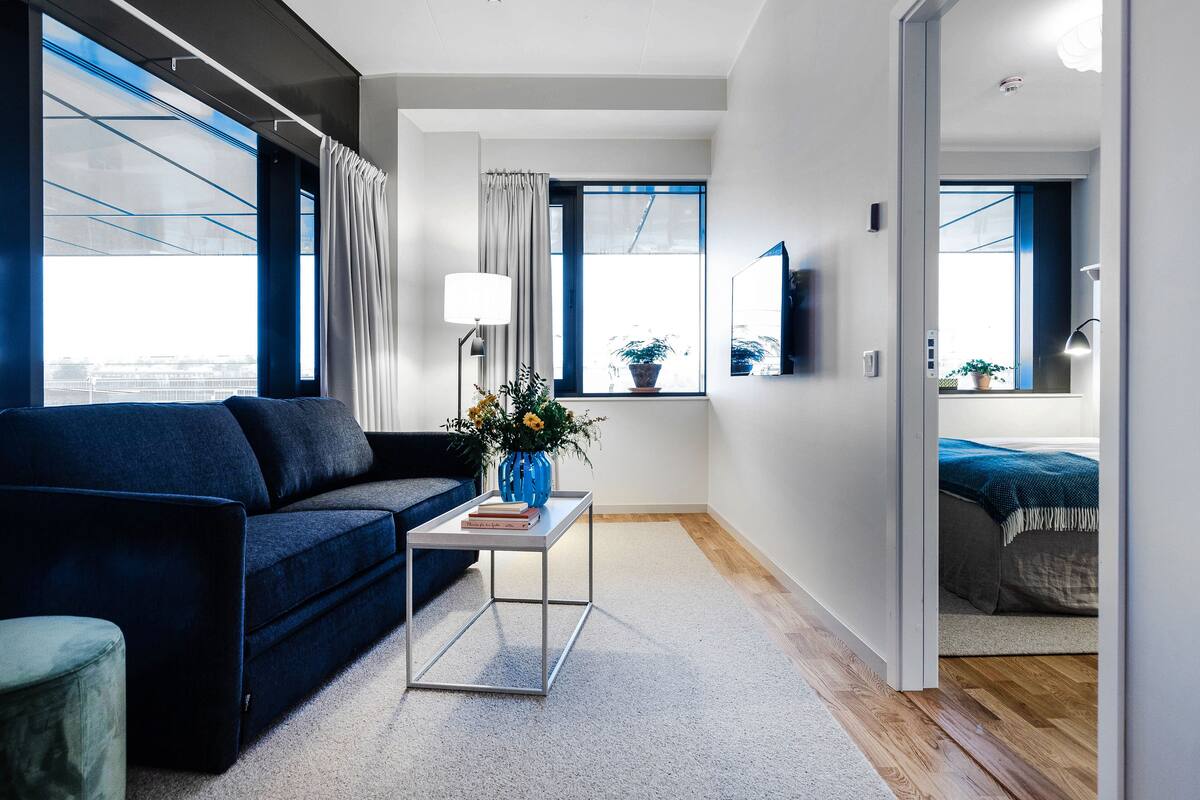 Una sala de estar moderna con un sofá azul oscuro, una mesita metálica con un florero y vistas a un paisaje urbano a través de amplios ventanales.