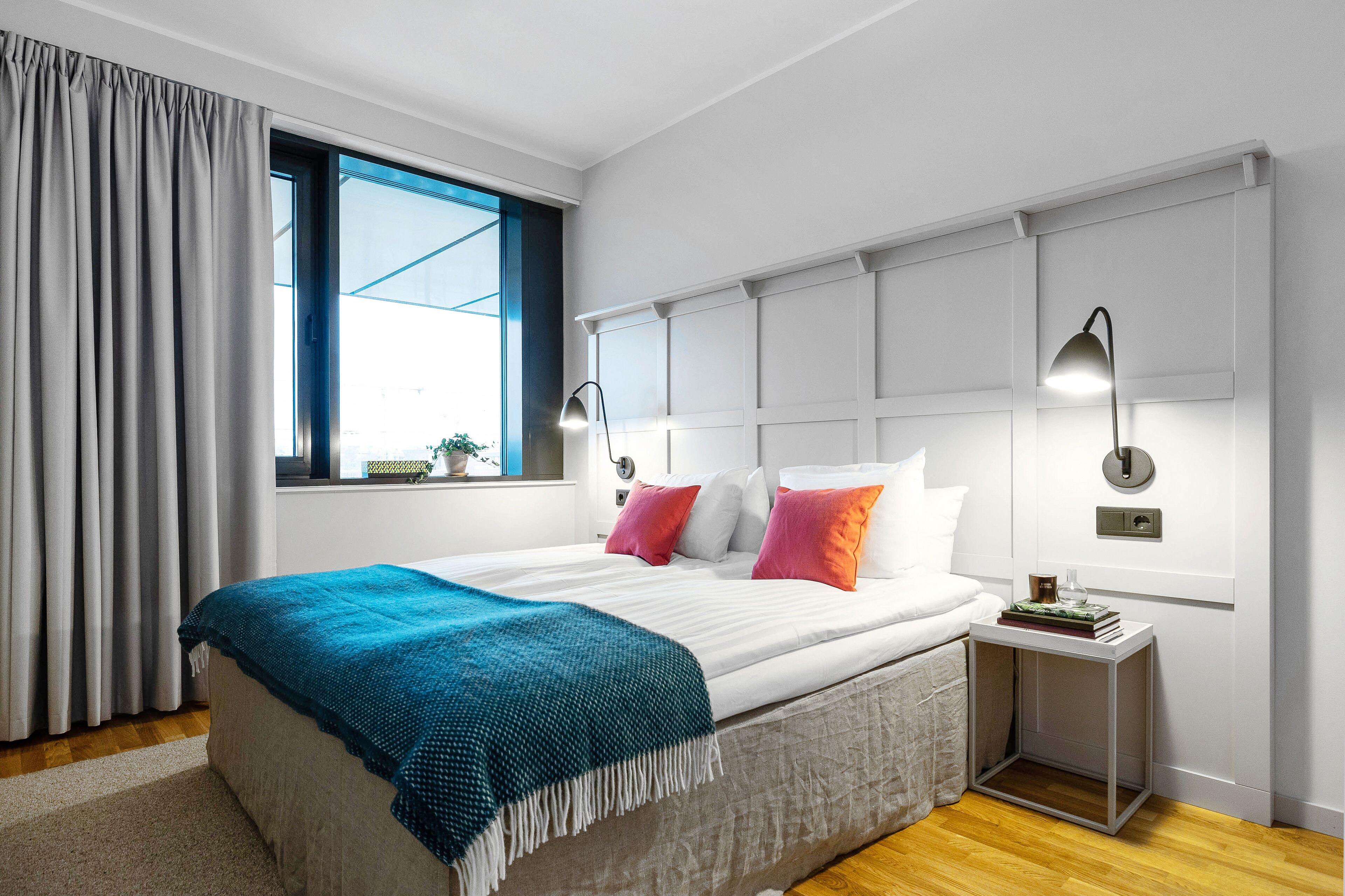 Ein modernes Schlafzimmer mit einem großen Bett, einem Fenster mit Vorhängen, Nachttischlampen und einem kleinen Tisch mit Büchern.