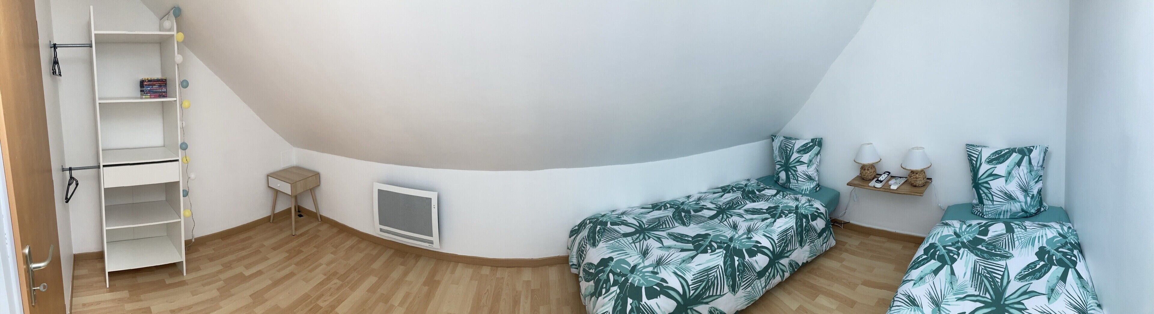 Een slaapkamer met een gebogen plafond, een bed met een tropisch bladmotief, een klein nachtkastje, een plank met decoratieve objecten, en een witte kledingkast met planken.