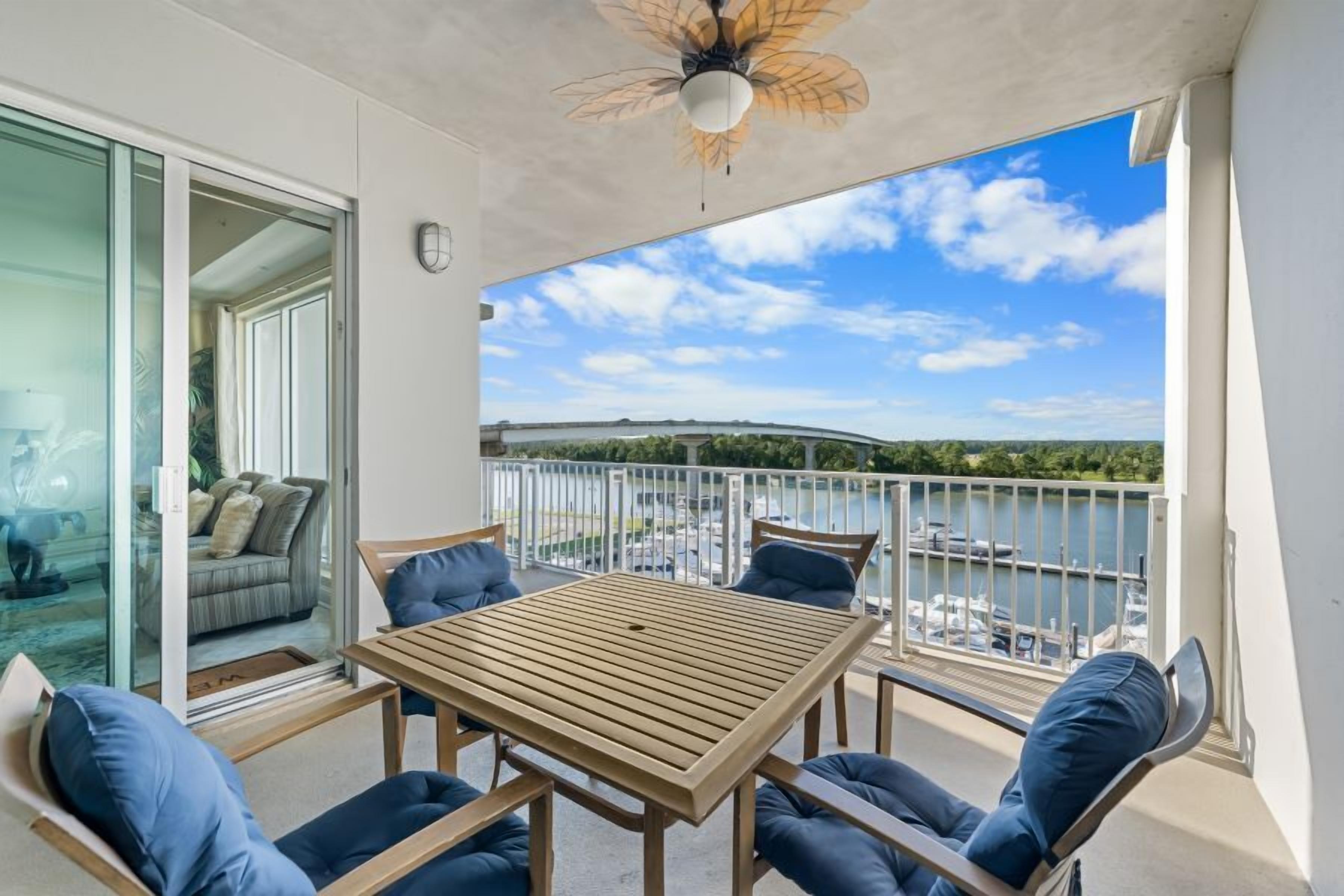 A balcony with a wooden table and chairs, a glass door leading to a room, and a view of a marina and bridge.