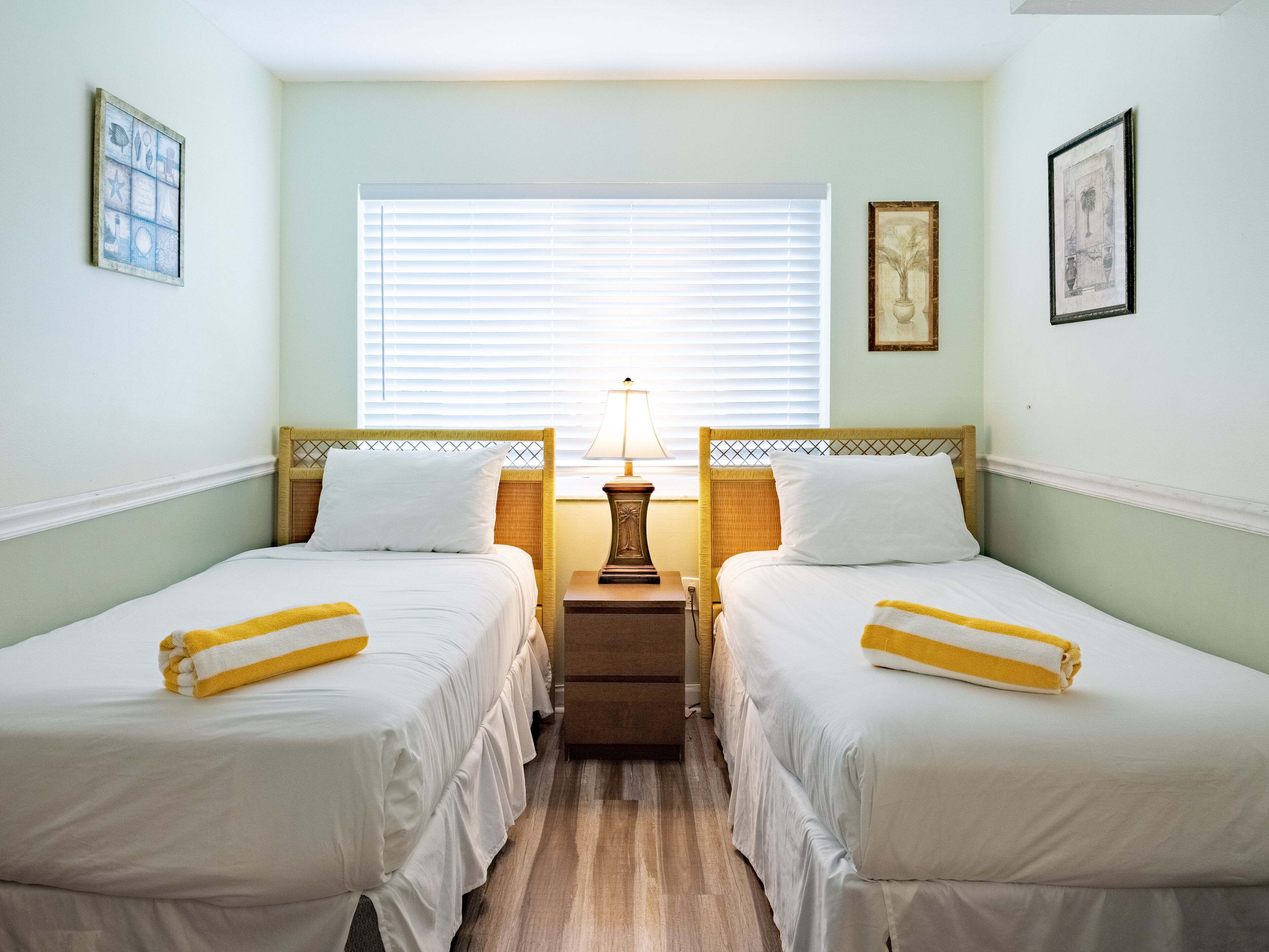 Una habitación de hotel con dos camas individuales, una mesita de noche con lámpara y una ventana con persianas.