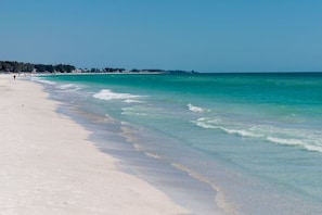 Een strand met helder turquoise water, een zandstrand en een verre boomlijn.